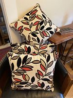 Set of three square pillows stacked in chair, showing abstract leaf design in cinnamon, green, grey with black velvet outlines.