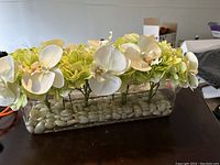 Side view of artificial flower arrangement with white orchids and green hydrangeas set in clear glass vase with white stones