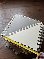 Stack of several interlocking foam play mats in light grey and off-white color on a wooden floor, showing measurement of one mat as approximately 22 inches square