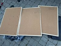 Three lightly used rectangular cork bulletin boards with wooden frames laid on ground, one board with multiple push pins attached.