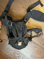 Black Ergobaby baby carrier with padded shoulder straps, adjustable buckles, and waist belt laid flat on wooden floor.