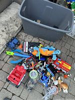 Plastic bin with assorted disassembled kids toys scattered around including vehicles, animals, action figure parts.