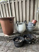 Six garden flowerpots including a large terracotta pot, three medium pots with hanging frames (white, grey-striped, terracotta), and two small black plastic pots on a stone patio near a wooden fence.
