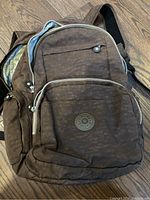Brown nylon Kipling backpack with zippered main compartment, front and side pockets, and adjustable shoulder strap shown against wooden floor