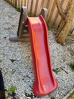 Side view of the Little Tikes children's slide showing red slide surface and brown support frame on gravel ground near fence and tree.