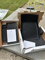 Photo of three black rubber floor mats for 2020+ Mercedes GLA250 in open boxes placed outdoors on a concrete surface. Instruction sheets visible on top of mats.