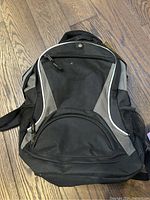 Photo of a new black and gray backpack on a wood floor, showing front view with zippered compartments and padded straps.