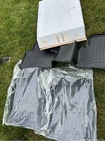Four black rubber floor mats for Mercedes-Benz GLS laid out on grass, with original white box behind them. Two mats are still inside clear plastic bags; two are outside the bags, showing channel designs and brand logo.