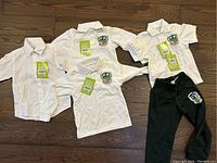White kid's school uniform clothing set on wooden floor including 2 long sleeve polos, 1 short sleeve polo, 1 dress shirt, 1 pair of black pants, all with school emblems and tags.