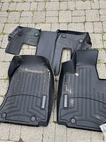 Three black floor mats arranged on stone pavement, two front mats with Mercedes-Benz metal logos, one rear middle mat without logo.