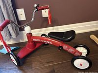 Red Radio Flyer balance bike with three wheels, black seat, metal frame and handlebar tassels.