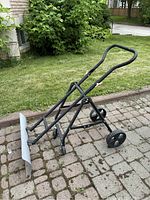 Full view of the used snow shovel on a stone patio, showing its metal blade, black metal frame with two plastic wheels, and foam grip handle.