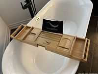 Photo shows a wooden bath table placed across a white bathtub. The table features multiple compartments including a wine glass holder, soap tray, and other sections, with extendable wooden sides. The brand 'Streamline' with a wave logo is visible embossed on the center compartment.