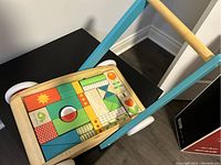 Photo shows a wooden push walker toy by Tender Leaf Toys with a brightly colored geometric shape sorter puzzle on the tray top. Handle painted teal blue and natural wood frame.