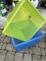 Two plastic rectangular storage boxes stacked, green on top and blue below, both without lids, photographed outdoors on paving stones.