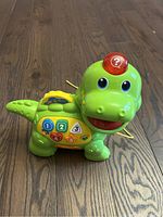 Green plastic dinosaur walking toy with buttons and no sound, on wood floor surface.