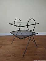 Front view of the two-tier end table showing glass top and metal mesh lower shelf with circular metal accents on sides.