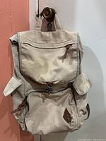 Full frontal view of beige canvas backpack with front flap, side pockets, and zippered main compartment.