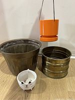 Four pots: one large dark metal pot, two vintage hammered brass pots, one orange hanging planter, and one small white ceramic cat face pot.