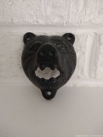 Front-facing view showing detailed face of bear-shaped cast iron bottle opener with mouth opening for removing bottle caps. Mounted on white painted brick background.