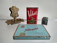 Front view of the vintage tobacco tins, silver-tone lighter, and brass ashtray showing items together with clear visibility of the Velvet and Player's tin logos and ashtray's ornate details.