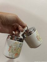 Showing two ceramic egg coddlers with floral patterns and metal lids with loop handles