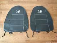 Two black Honda-branded universal seat cover pads lying on wooden floor, showing front side with logo and straps.