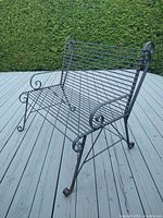 Full view of the vintage wrought iron bench showing the slatted seat and backrest, scrollwork armrests, and sturdy legs on a gray wooden deck with hedge background.