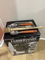 Photo showing various utensils stacked on original Farberware box including wooden honey dipper and orange handled spoons.