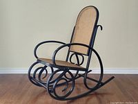 Front-left angled view of vintage black bentwood rocking chair with cane seat and backrest, showing entire chair and wood floor.