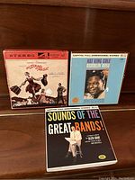 Three vintage reel-to-reel tape boxes on a wooden surface, showing album covers for The Sound of Music, Nat King Cole Ramblin' Rose, and Sounds of the Great Bands