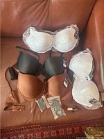 Five padded underwire bras arranged on a couch. Three white bras wrapped in plastic, one black bra with tags, one brown bra with tags. Small plastic packages containing bra accessories in view.