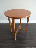Full front view of round teak side table on dark floor, showing table legs and top surface.