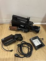 Front and top view of JVC Saticon video camera with wooden handle, battery pack, JVC accessory and power cable on floor