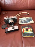 Four items on brown leather sofa showing Yashica Electro 35 camera, Kodak compact camera, Yashica manual and Kodak Master Photoguide booklet