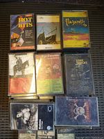 Nine vintage cassette tapes displayed on a black textured surface, featuring artists such as Nazareth, Elvis Presley, Fleetwood Mac, and Star Tracks. Cases and cover art show signs of wear and dust.