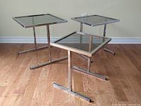 Three chrome steel framed smoked glass square nesting end tables of different sizes shown on hardwood floor against wall.