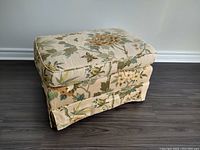 Front and left side view of vintage rectangular ottoman with floral fabric upholstery on beige background showing faded spots and wheel underneath