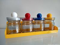 Front view of the set showing four clear plastic jars with lids in white, red, blue, and yellow knob colors arranged in yellow wall mount bracket.