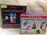 Photo showing two kids craft kits. The first is the 'Lava Lip Gloss Kit' by h2o naturals with a damaged box. The second is the 'Enchanting Craft Kit' by Kid Made Modern with colorful character illustrations on the box and some visible wear.