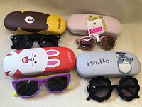 Top-down view of the four children's sunglasses placed above their cartoon-themed hard cases on a beige surface.