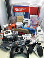 Wide view of the entire lot showing various new electronics items including gaming controller, Bluetooth earbuds, storage cases, and boxed accessories.