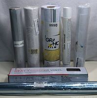 Six rolls of heat transfer vinyl standing upright against a wall, one roll in a box labeled Heat Transfer Vinyl with instructions.