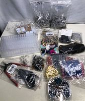 Overview of multiple clear plastic bags containing metal charms, beads, and findings, alongside a plastic organizer box and some packaged pliers.