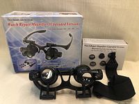 Box and contents for watch repair magnifier kit showing the product box with product image and text along with a smaller storage box and black head strap.