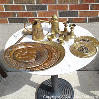 Photo showing large assortment of brass and copper items on a round table including plates, watering can, candle holders, cups, and wall hanger.