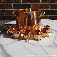 Front view of the copper pot with lid and 7 small barrel-shaped copper cups with brass handles arranged in front.