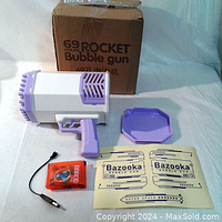 Photo of the 69 hole rocket bubble gun with accessories including purple bubble tray, battery pack, charging cable, two stickers, and original cardboard box.