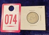 Image of the 1968 Canadian quarter in a cardboard holder next to a tag with the number 074.
