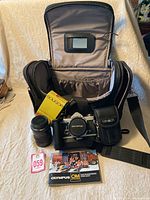 Open deluxe camera bag with Olympus OM System camera, lenses, Minolta accessory case, and Olympus catalog booklet arranged in front of the bag.